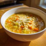 A close-up of a white bowl filled with warm pastina soup, featuring carrots, celery, and topped with grated Parmesan cheese.
