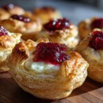 Close-up of golden, flaky pastry cups filled with melted brie and topped with bright red cranberry sauce, featuring cranberry brie bites.