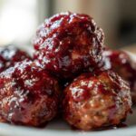 A close-up shot of several glossy, saucy cranberry meatballs piled on a white plate.