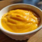 Close-up of smooth, bright orange beer cheese dip served in a small white bowl on a wooden surface.