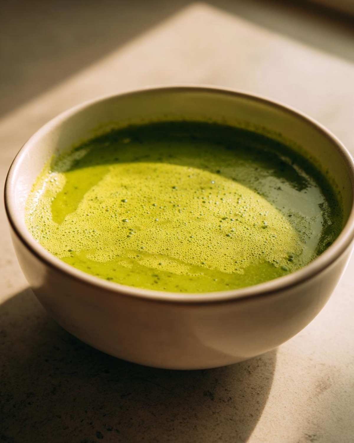 Close-up of a bowl of vibrant, creamy celery soup with a frothy top, illuminated by natural light.