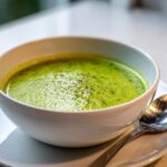 Close-up of a bowl of vibrant green, creamy celery soup, topped with cracked black pepper.