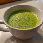 Close-up of bright green, foamy celery soup served hot in a white mug.