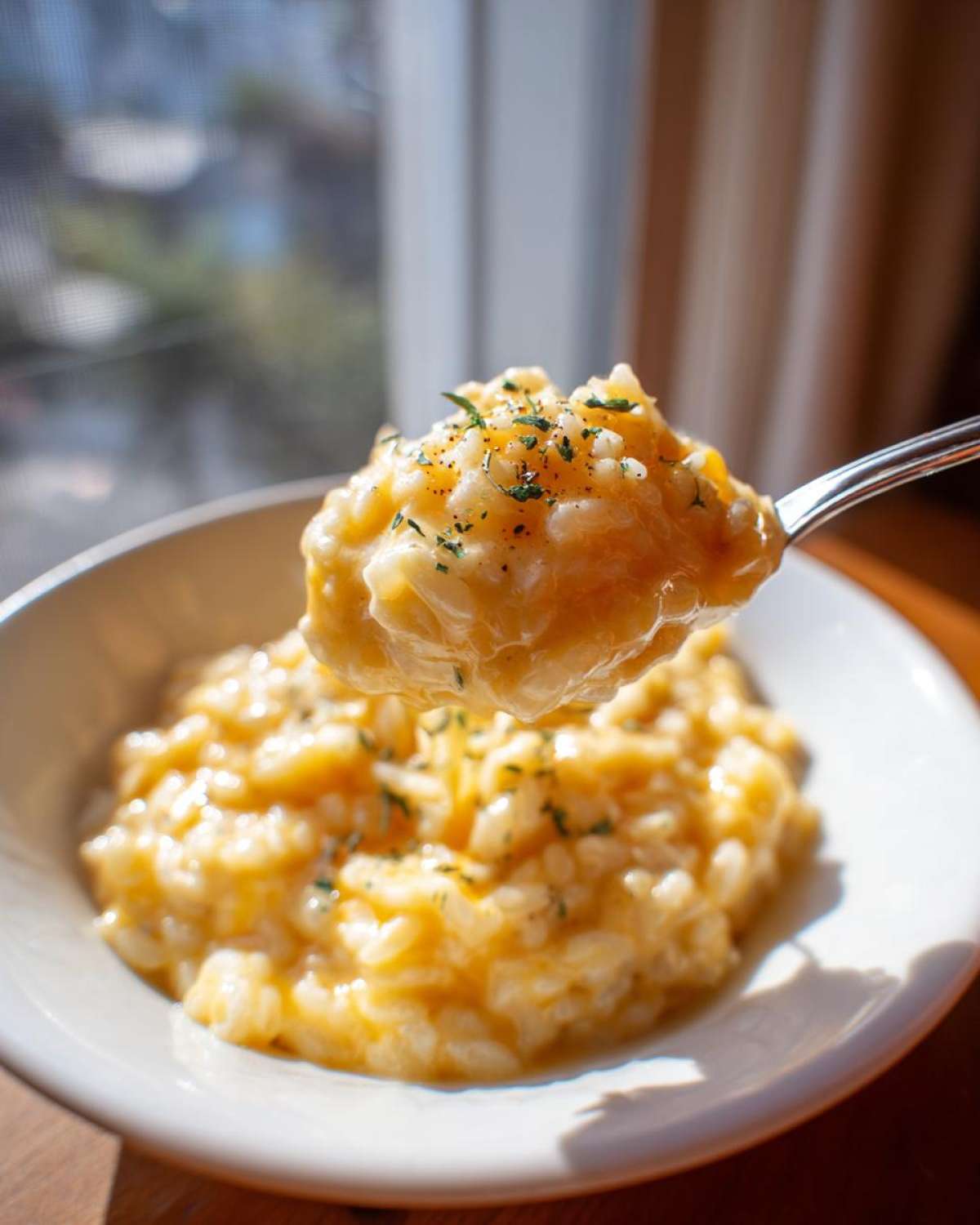 A spoonful of creamy, bright yellow cheesy rice garnished with herbs, lifted from a white bowl.