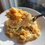 A spoonful of creamy, melted cheesy rice being lifted above a white bowl, showing melted cheddar.