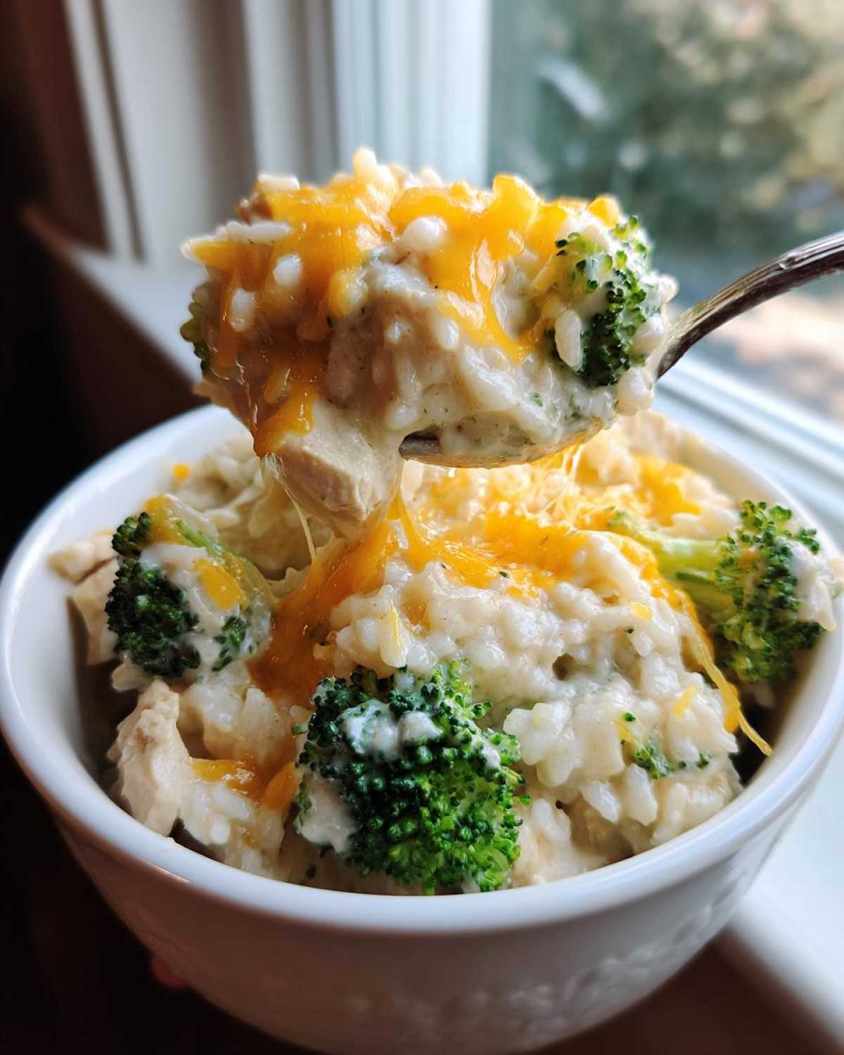 A spoonful of creamy chicken broccoli rice casserole topped with melted cheddar cheese being lifted from a white bowl.