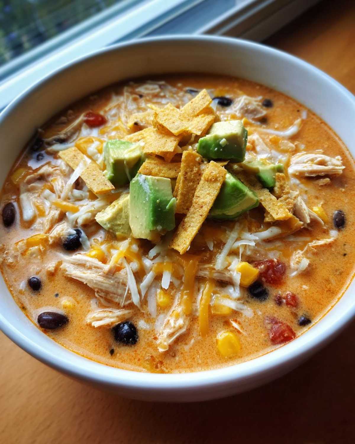 A close-up of a bowl of creamy chicken tortilla soup topped with shredded cheese, avocado, and tortilla strips.