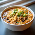 A white bowl filled with creamy chicken tortilla soup, topped with shredded cheese, avocado, and tortilla strips.