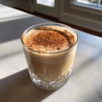 A glass of creamy coquito topped generously with ground cinnamon, sitting on a sunlit marble counter.