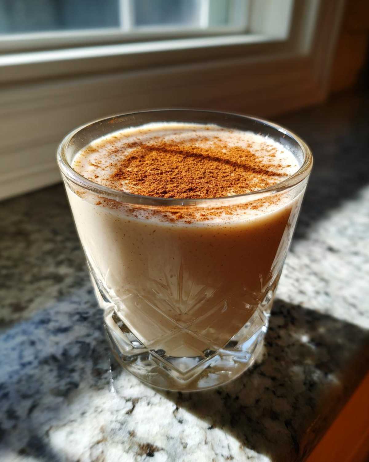 A glass of creamy coquito topped generously with ground cinnamon, catching sunlight.