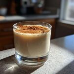 A glass of creamy coquito topped generously with ground cinnamon spice, sitting on a sunlit countertop.