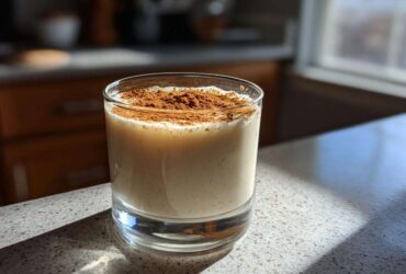 A glass of creamy coquito topped generously with ground cinnamon spice, sitting on a sunlit countertop.