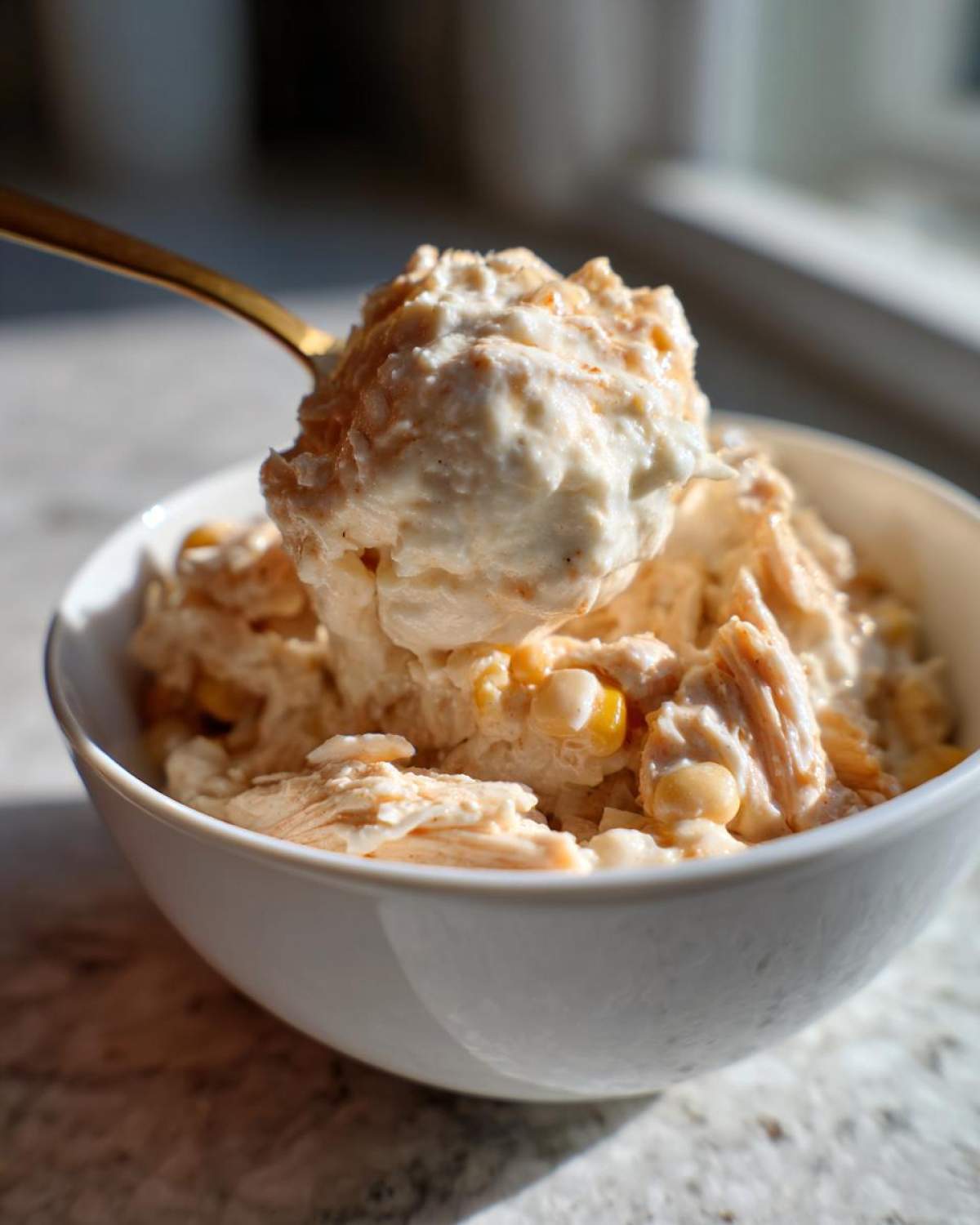 A golden spoon lifts a creamy scoop of crockpot white chicken chili, showing shredded chicken and corn kernels.