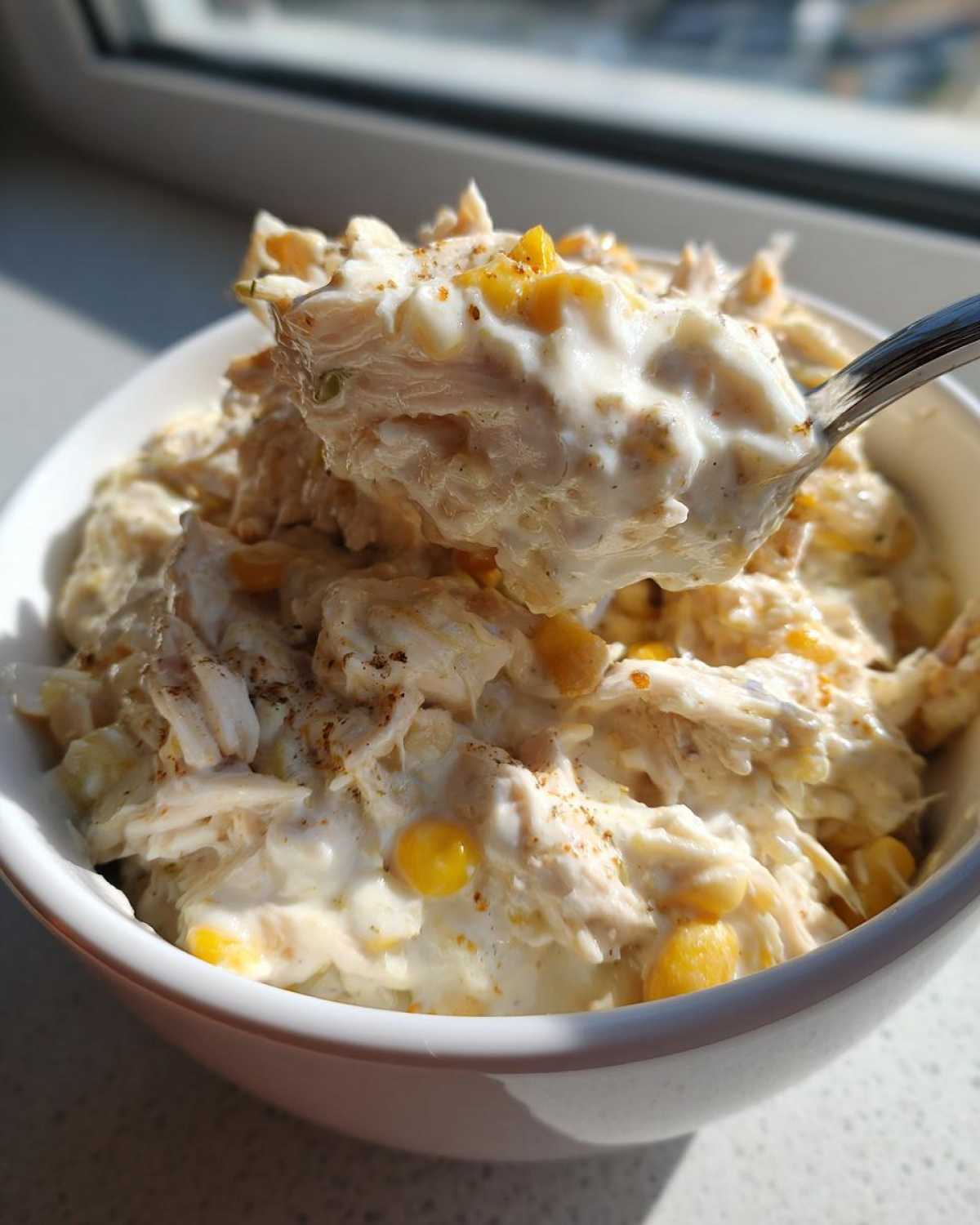 A spoonful of creamy crockpot white chicken chili, featuring shredded chicken and corn kernels, being lifted from a white bowl.