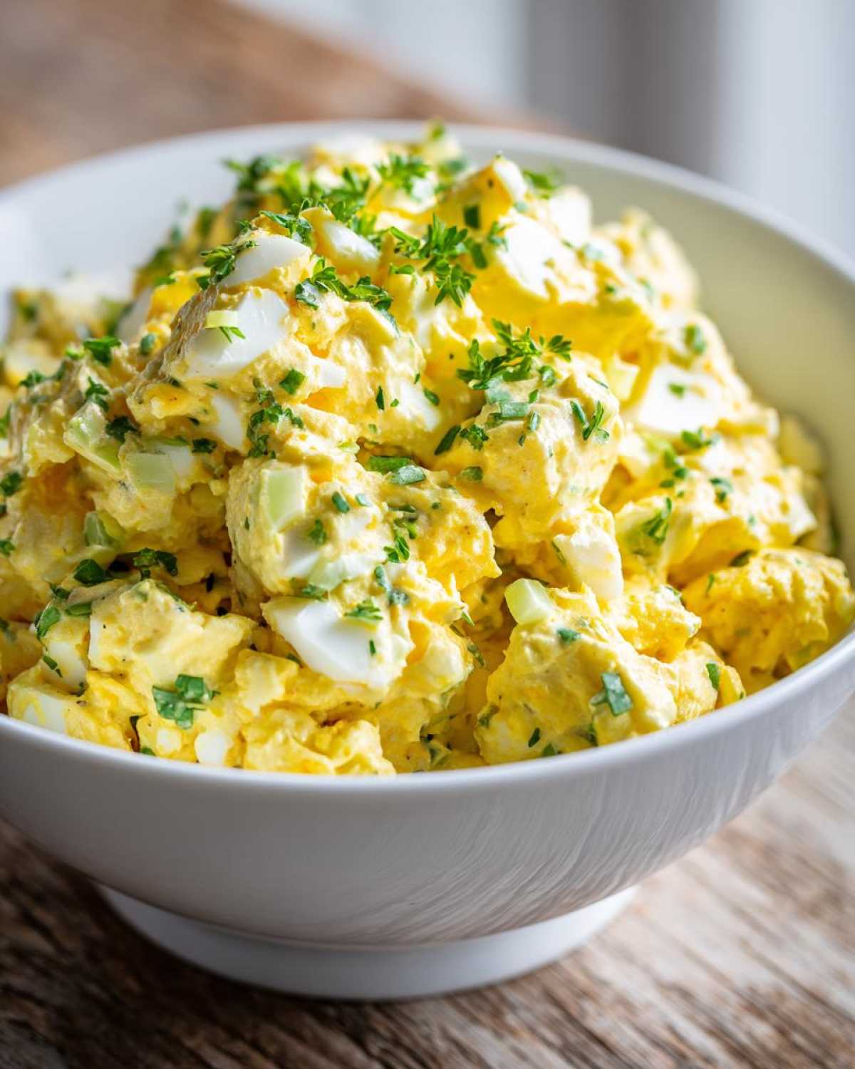Close-up of a white bowl filled with creamy, yellow egg salad recipe, topped with fresh chopped parsley.