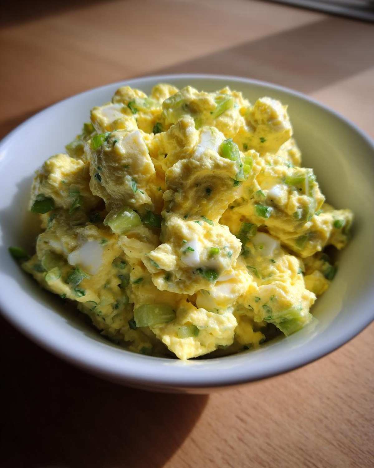 A close-up view of creamy, yellow egg salad recipe mixed with chopped green celery and herbs, served in a white bowl.