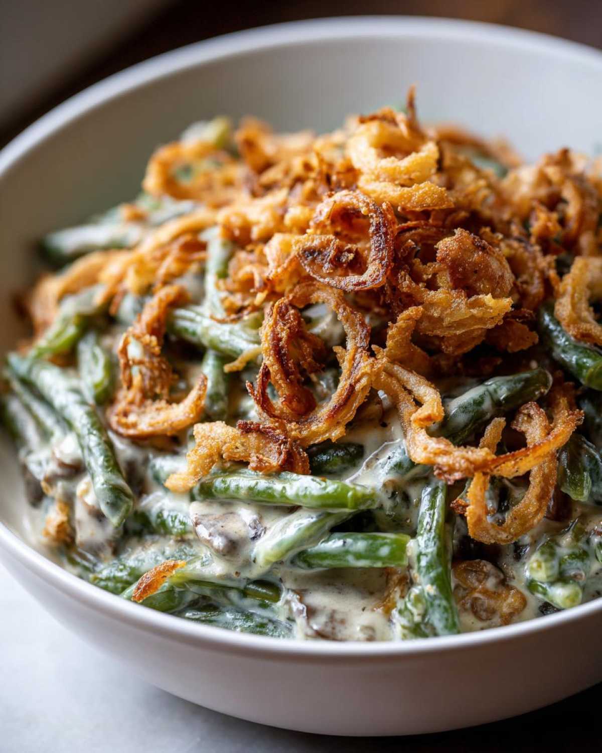 A close-up of a white bowl filled with creamy green bean casserole topped generously with crispy fried onions.
