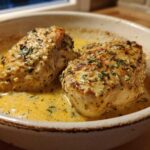 Two golden-brown chicken breasts simmering in a rich, yellow creamy lemon chicken sauce, garnished with herbs.