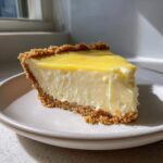 A thick slice of creamy lemon pie with a bright yellow topping and crumbly graham cracker crust on a light plate.