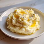 A serving of fluffy, creamy mashed potatoes topped with pools of melted butter and cracked black pepper.