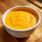 Close-up of vibrant orange, creamy nacho cheese sauce sprinkled with spices in a small white bowl.