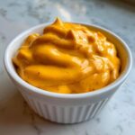 Close-up of thick, vibrant orange nacho cheese sauce swirled in a small white bowl.