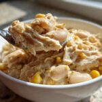 A spoonful of creamy crockpot white chicken chili, showing shredded chicken, white beans, and corn, lifted from a white bowl.