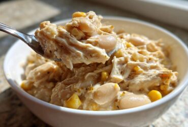 A spoonful of creamy crockpot white chicken chili, showing shredded chicken, white beans, and corn, lifted from a white bowl.