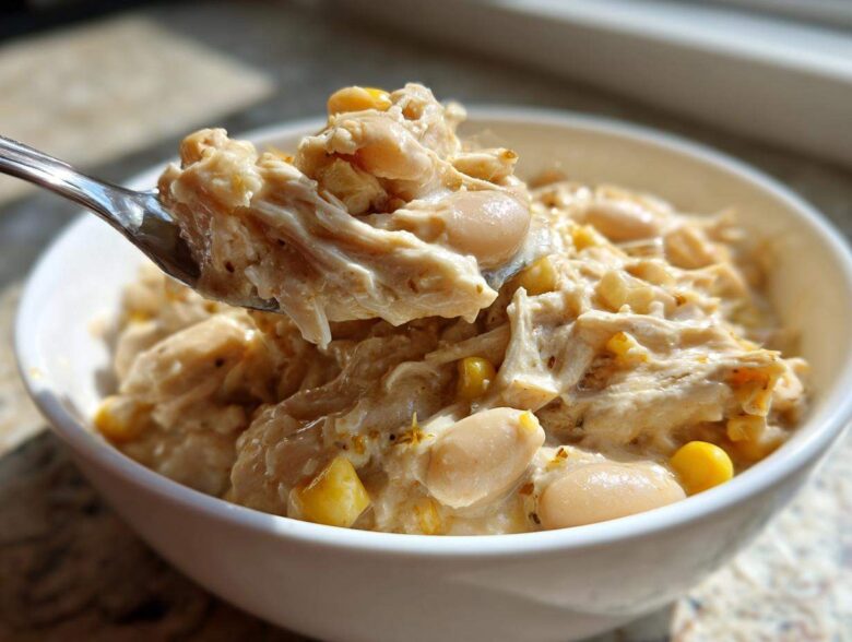 A spoonful of creamy crockpot white chicken chili, showing shredded chicken, white beans, and corn, lifted from a white bowl.