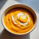 A vibrant orange squash soup topped with a white cream swirl in a light-colored bowl.