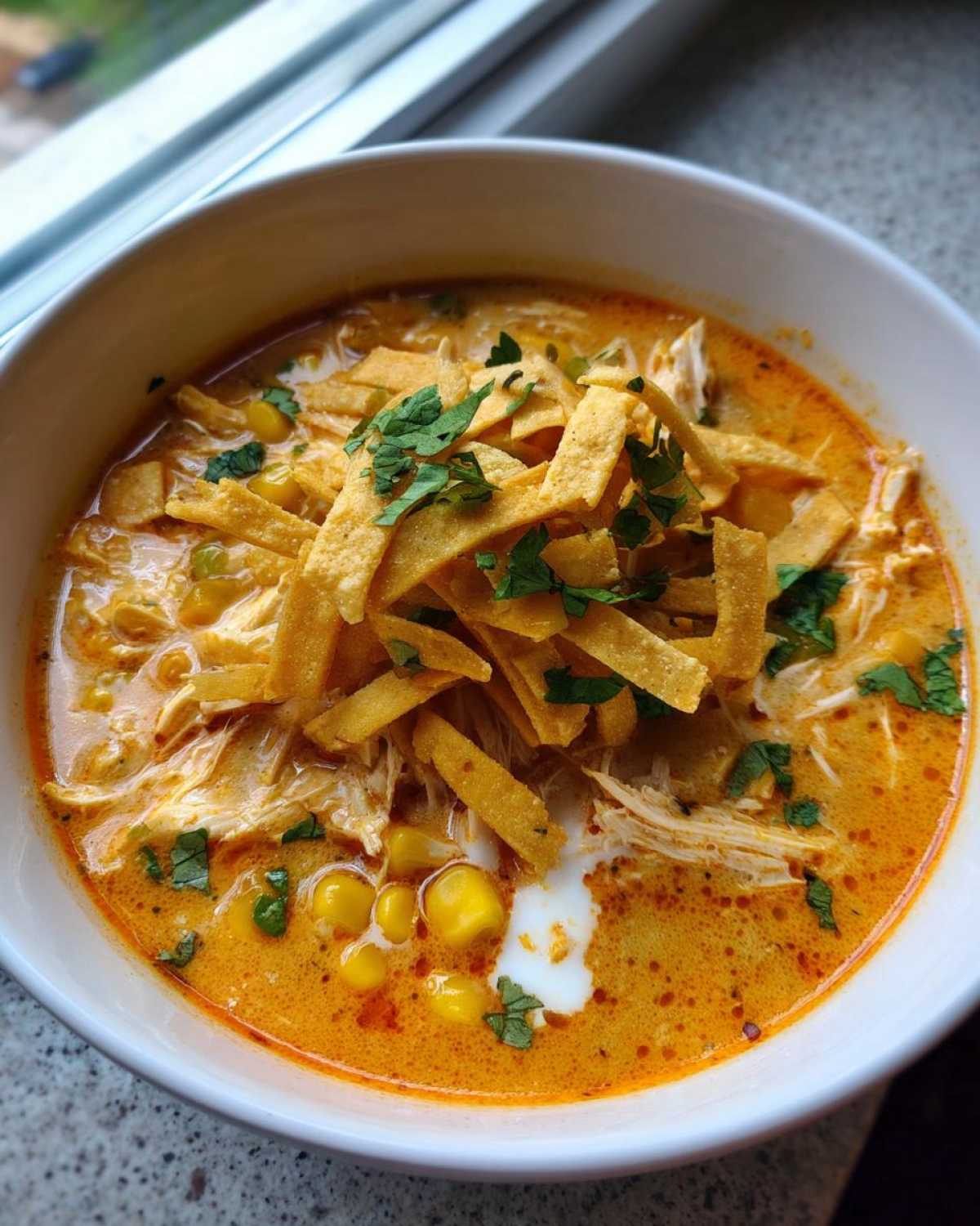 A close-up of creamy, orange-hued tortilla soup topped with shredded chicken, corn, tortilla strips, and cilantro.