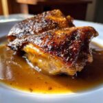 Two pieces of perfectly cooked braised chicken thighs with crispy, dark skin resting in a rich brown sauce on a white plate.
