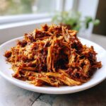 A close-up of tender, saucy crock pot pulled pork piled high on a white plate.
