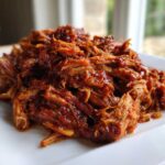A close-up of moist, saucy crock pot pulled pork piled high on a white square plate.