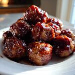 A close-up of several Crockpot grape jelly meatballs piled high on a white plate, glistening with sauce.