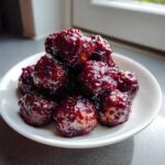 A close-up of several shiny, dark red Crockpot grape jelly meatballs piled on a small white plate.
