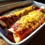 Close-up of two easy chicken enchiladas covered in red sauce and melted cheddar cheese in a white baking dish.