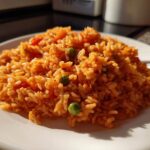A close-up of a serving of bright orange Spanish rice with visible peas and tomato pieces on a white plate.