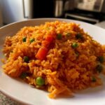 A close-up of a mound of vibrant orange spanish rice mixed with bright green peas and diced carrots on a white plate.