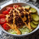A vibrant cheeseburger bowl featuring seasoned ground beef, shredded lettuce, cherry tomatoes, pickles, and a drizzle of special sauce.