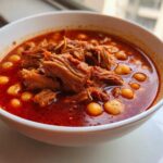 Close-up of a white bowl filled with vibrant red Pozole rojo, featuring shredded meat and hominy kernels.