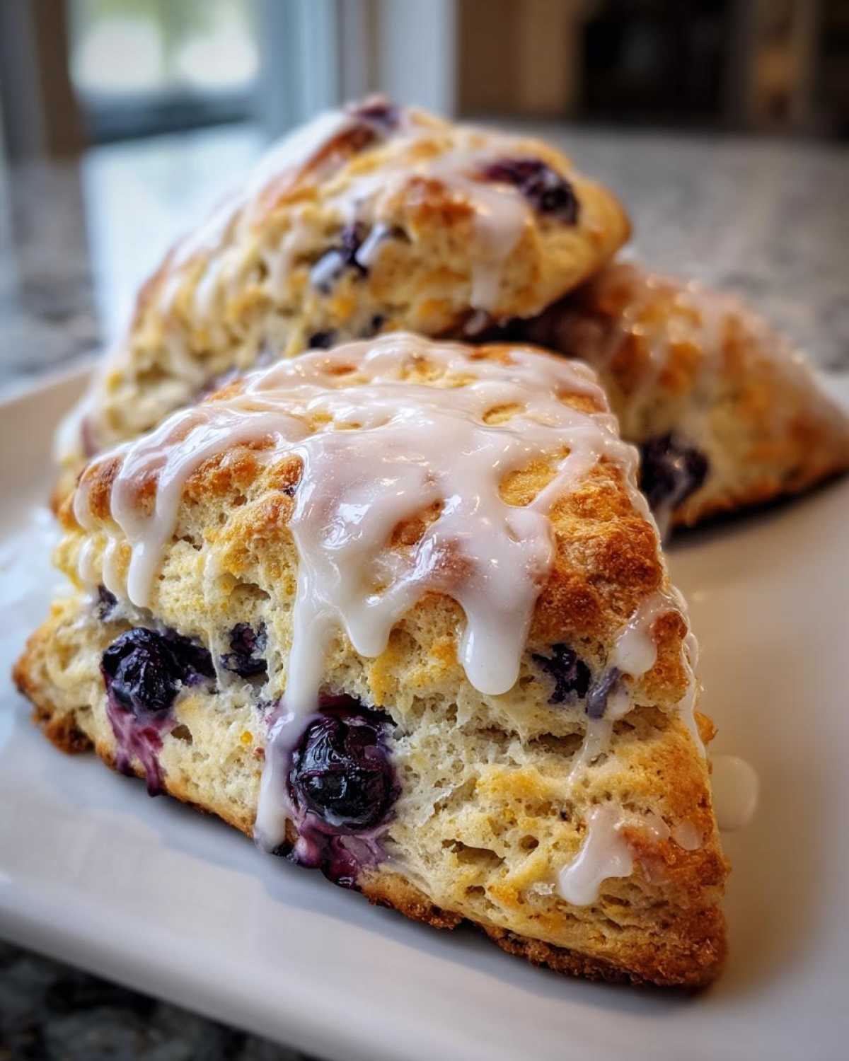 Close-up of a tall, flaky blueberry scone drizzled heavily with white vanilla glaze.