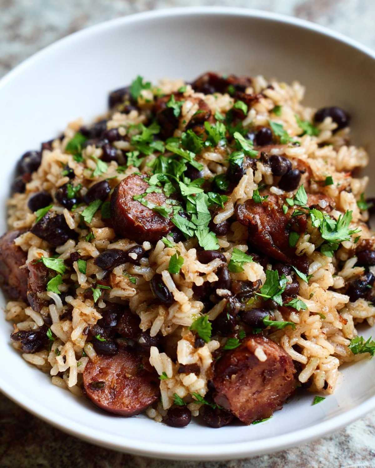 Close-up of a white bowl filled with savory black beans and rice mixed with sliced sausage and topped with fresh cilantro.