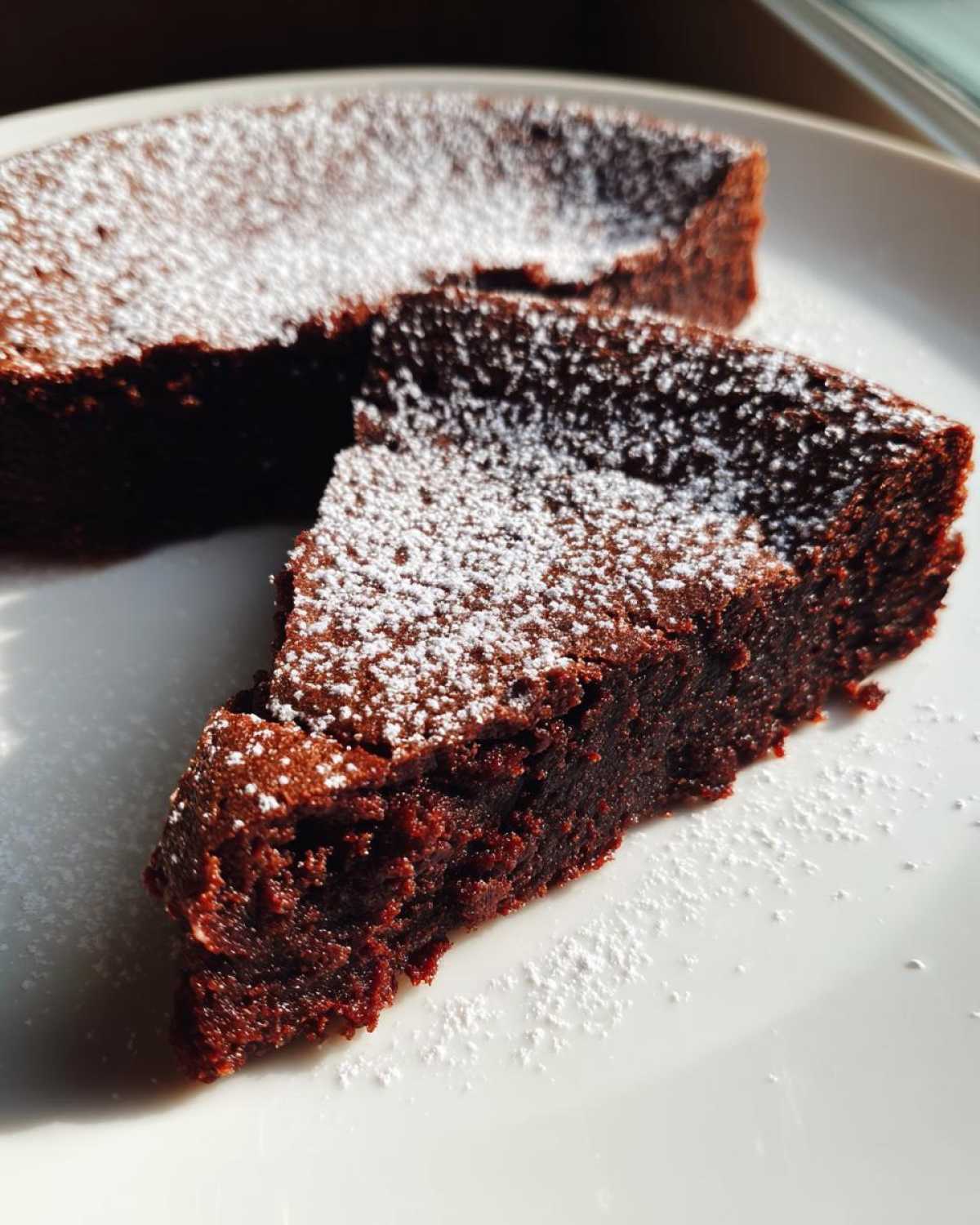 A close-up of a rich, dark slice of flourless chocolate torte dusted generously with powdered sugar on a white plate.