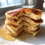 Close-up of a tall stack of fluffy almond flour pancakes with a bite taken out, drizzled with maple syrup.