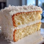 Close-up of a fluffy slice of coconut cake layered with white frosting and covered in shredded coconut.