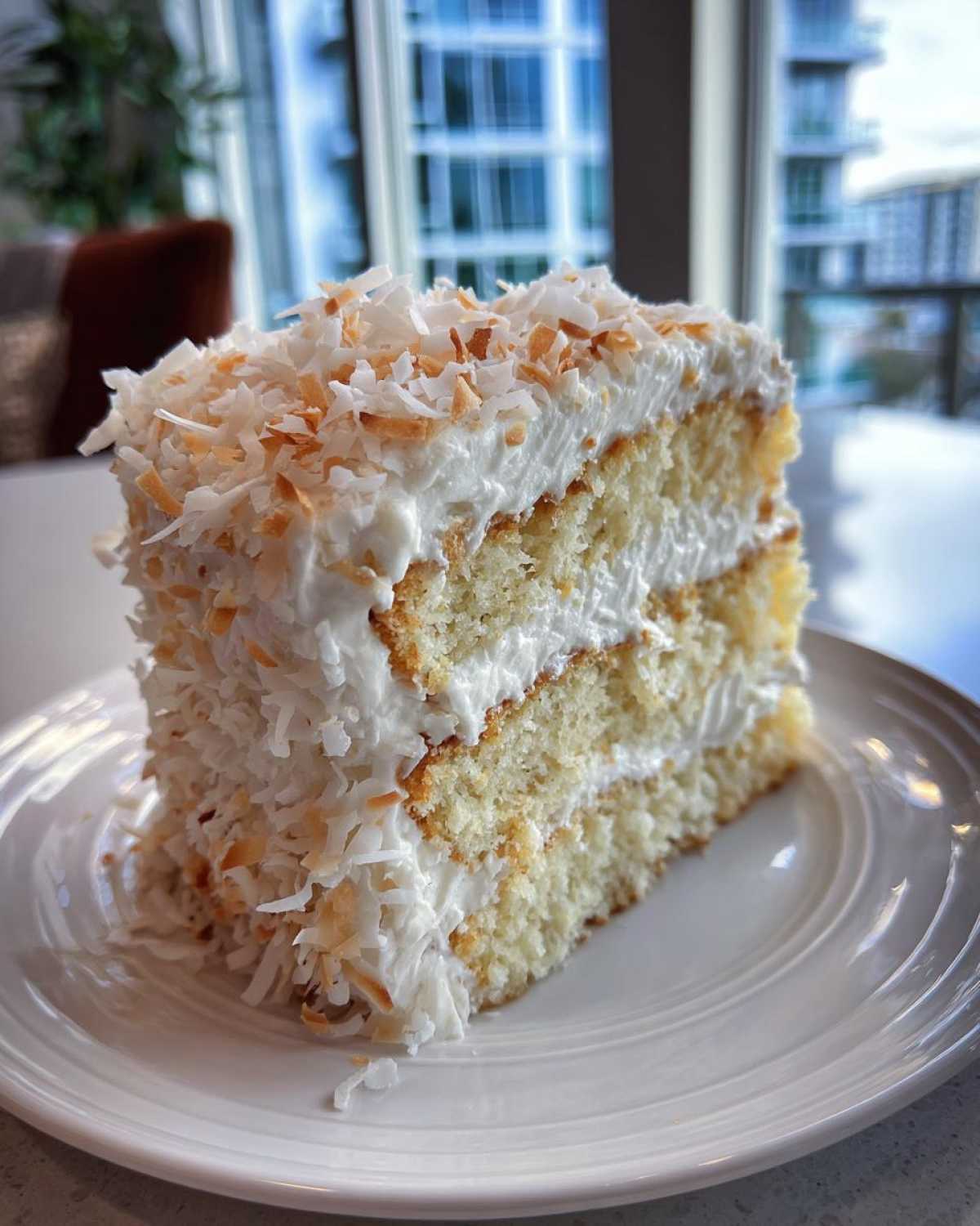 A tall slice of fluffy coconut cake with white frosting and toasted coconut flakes on a white plate.