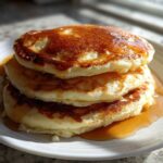 A stack of three fluffy pancakes drizzled generously with golden syrup, sitting on a white plate.
