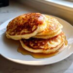 A stack of three golden brown, fluffy pancakes generously drizzled with maple syrup on a white plate.