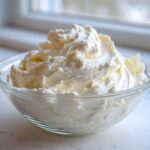 A clear glass bowl filled with perfectly swirled, creamy vanilla buttercream frosting, set near a bright window.